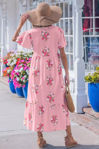 Woman in a pink floral dress with a sun hat, standing outdoors near potted plants.
