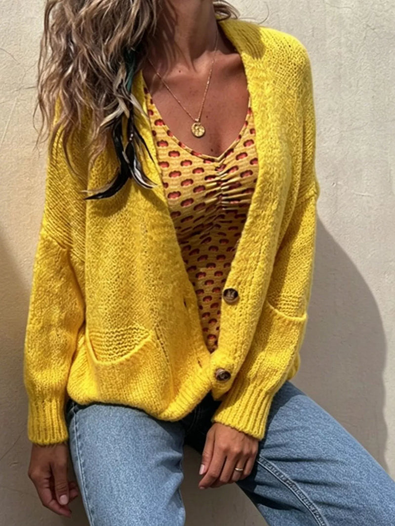 A woman wearing a bright yellow chunky knit cardigan over a yellow and red patterned camisole, paired with light blue jeans, leaning against a textured beige wall.