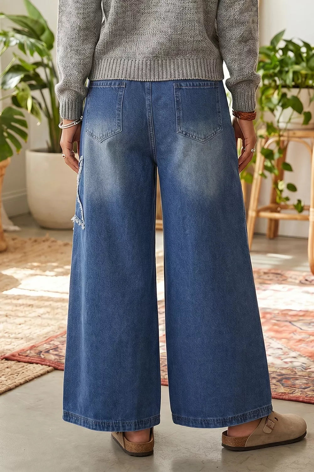 A person shown from the back wearing wide-leg blue denim jeans with a faded wash, a gray knit sweater, and tan slip-on shoes in a bright indoor room with plants and rugs.