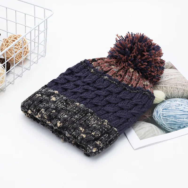 Knitted beanie with pom-pom on a white surface with yarn balls and a basket in the background.