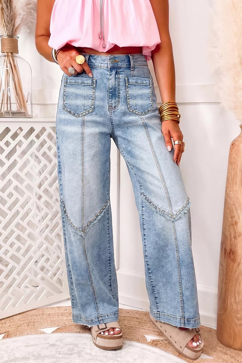 A person wearing light-wash blue wide-leg denim jeans with decorative contrast stitching and zippered patch pockets, styled with a pink sleeveless top and beige platform sandals in an indoor setting.