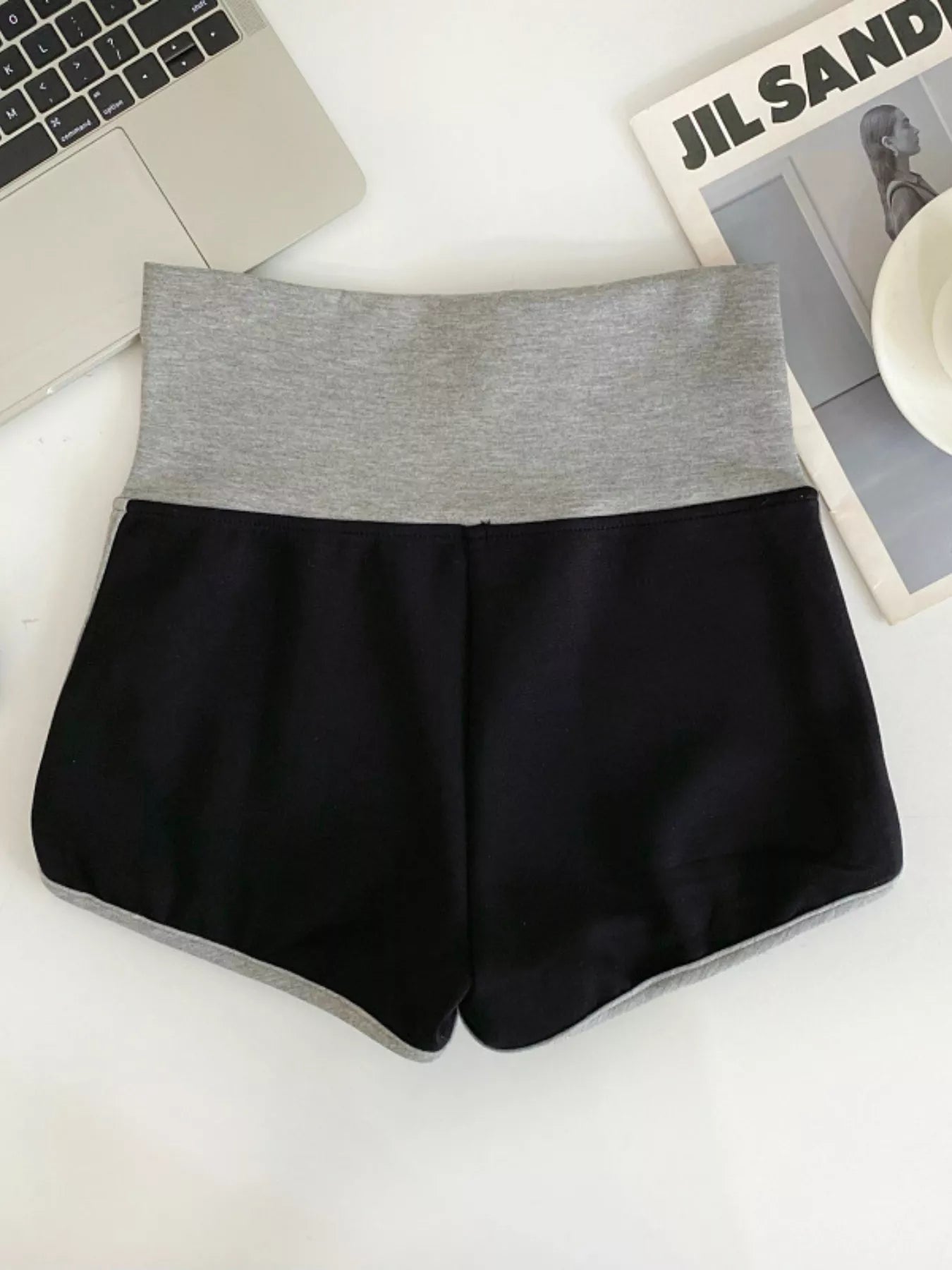Black shorts with gray waistband on a white surface with a laptop and magazine in the background.