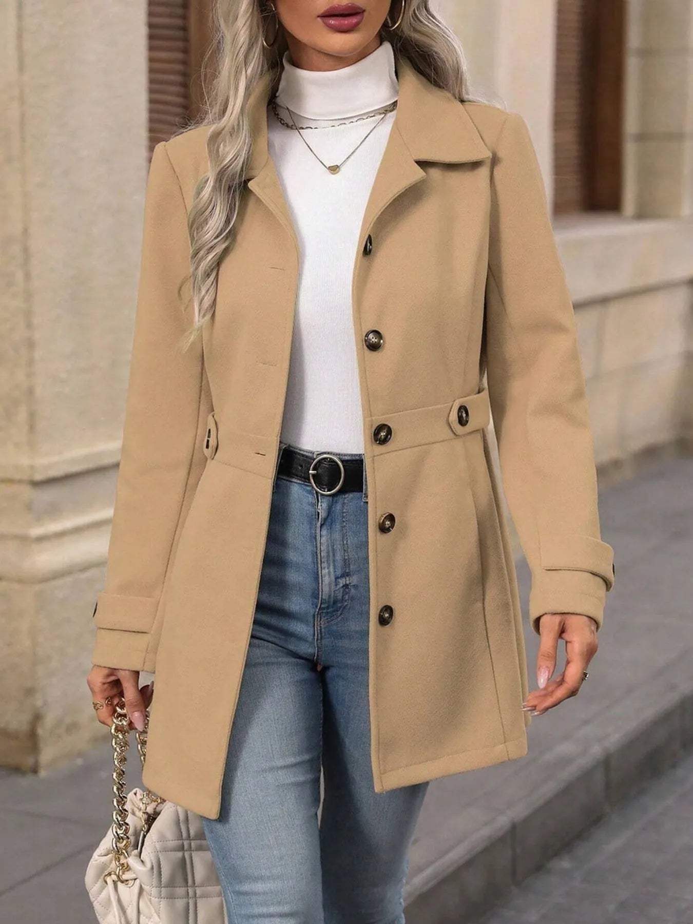 Woman wearing a beige coat, white turtleneck, and blue jeans outdoors.