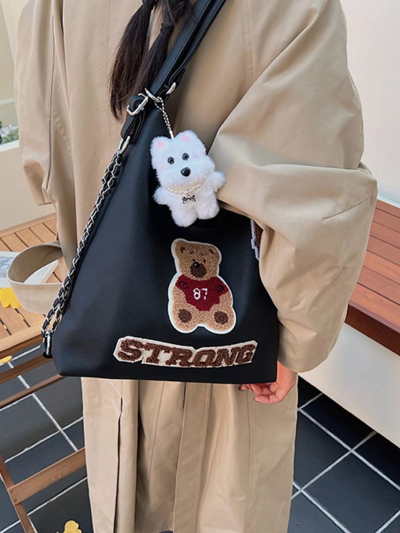 Person holding a black bag with teddy bear and dog design, wearing a beige coat.