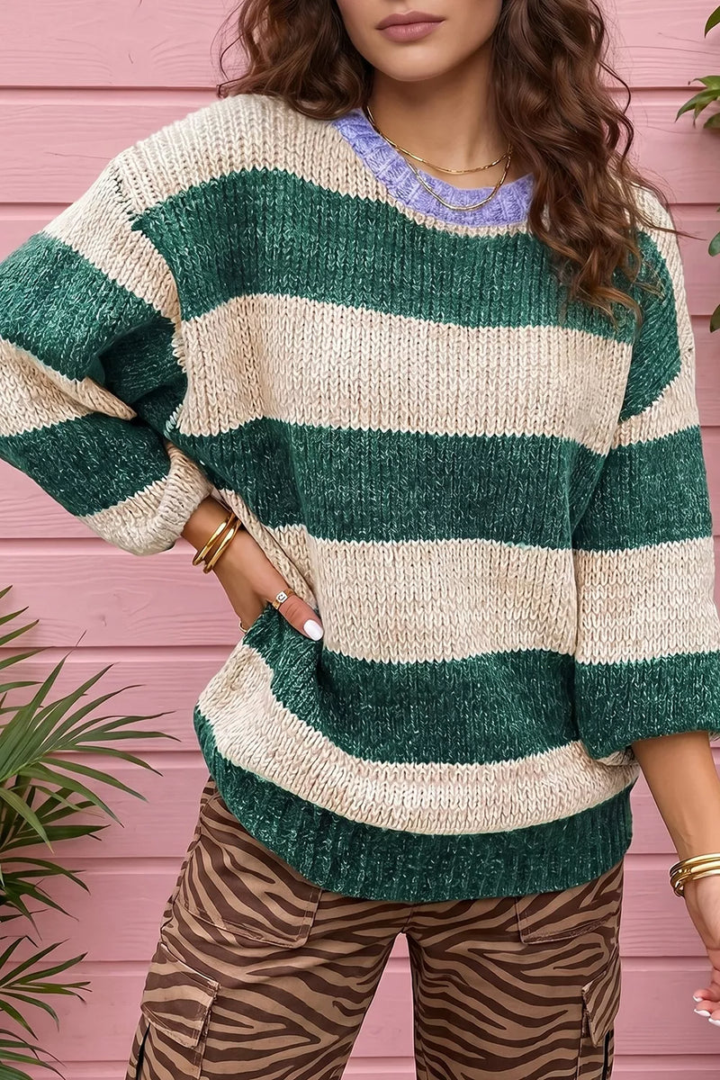 A woman wearing a chunky knit sweater with horizontal stripes in forest green and cream, paired with brown and black zebra-print pants, standing against a pink wooden wall.