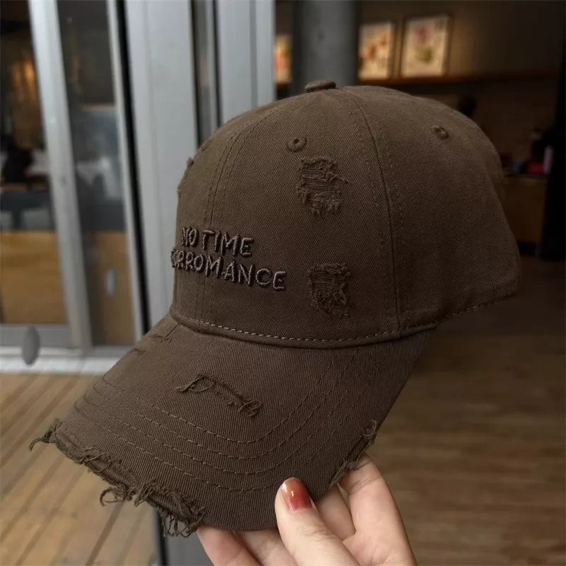 Brown distressed cap with 'No Time For Romance' text held by a hand indoors.