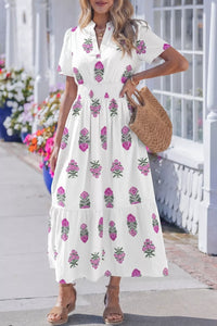 Woman wearing a white dress with pink floral patterns, standing on a sidewalk with flowers in the background.