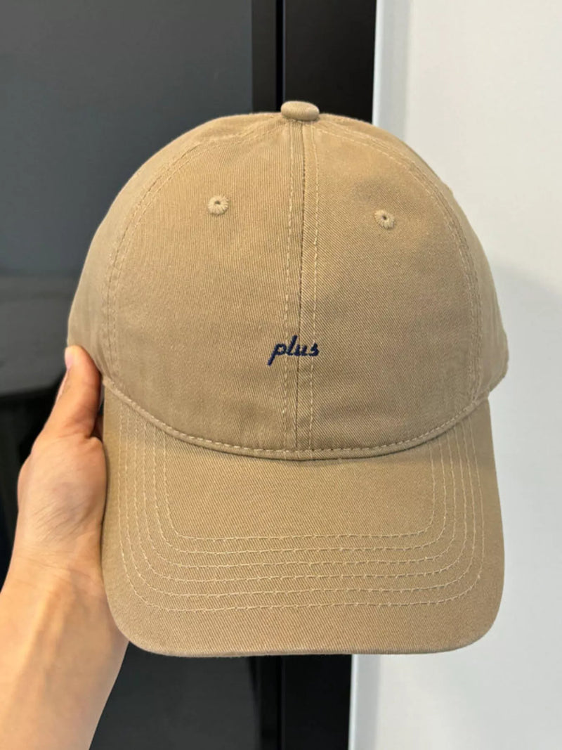 Beige cap with 'plus' embroidery held by a hand against a neutral background