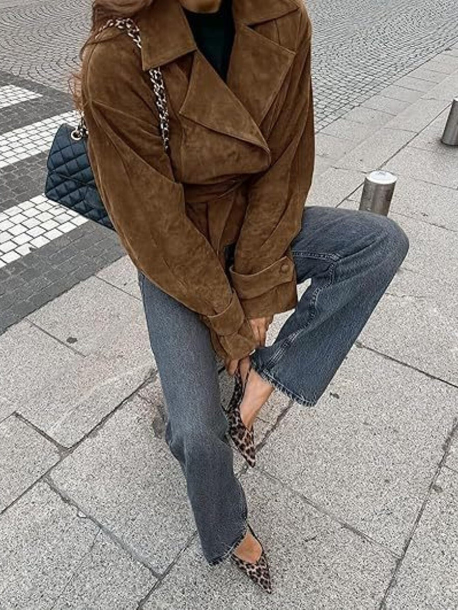 Person wearing a brown suede jacket and blue jeans on a city street.