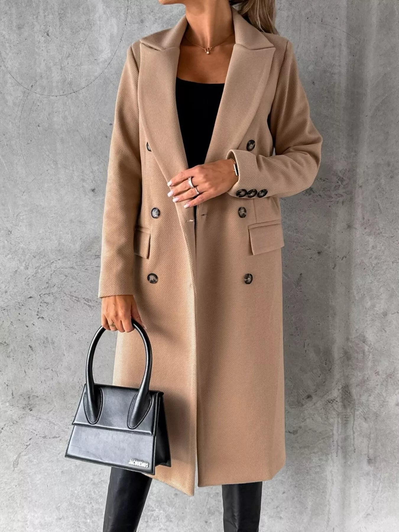 Person wearing a beige coat holding a black handbag against a gray concrete wall.