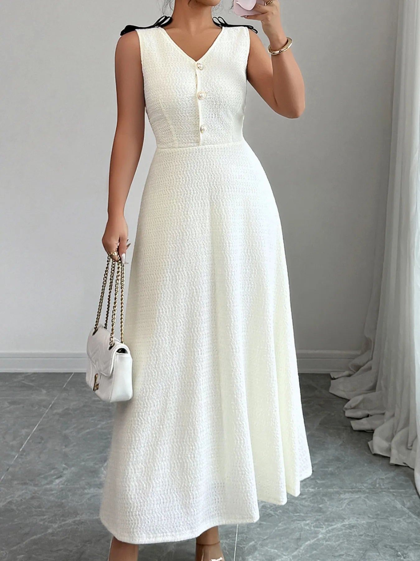 Woman wearing a white sleeveless dress holding a white handbag indoors.