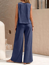 Woman wearing a blue sleeveless top and wide-leg pants standing against a beige wall.