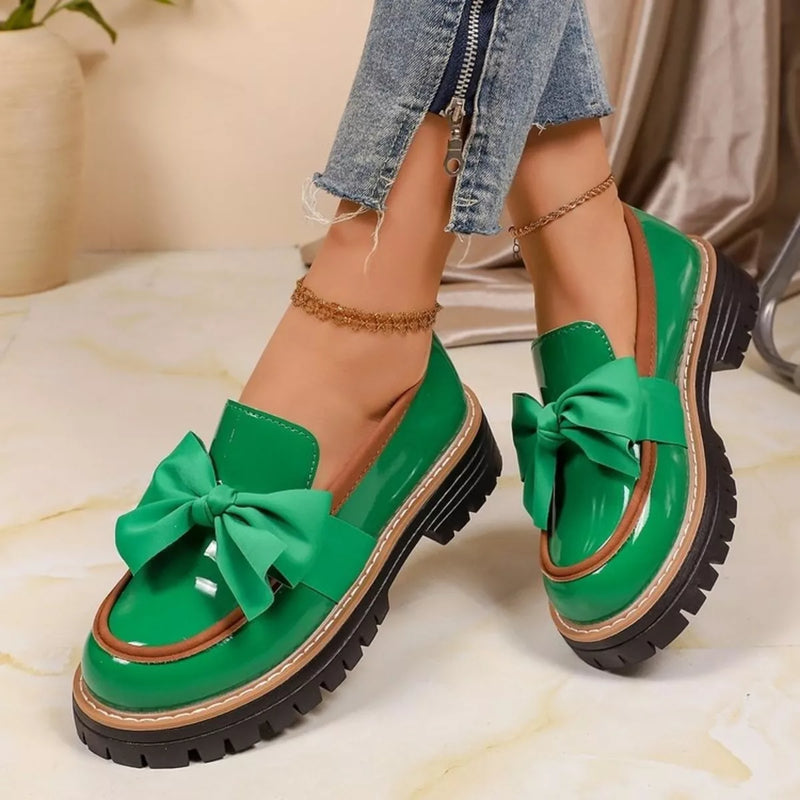 A pair of bright green patent leather slip-on loafers with large satin bows and chunky black platform soles, shown on a model's feet wearing frayed blue jeans.