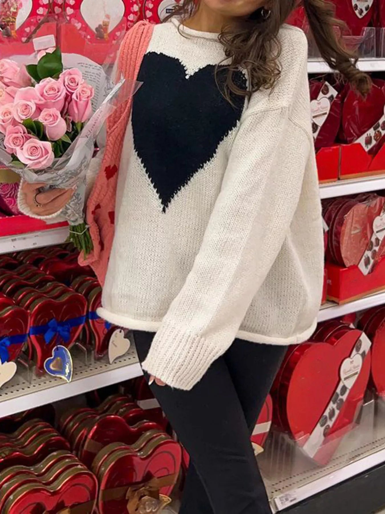 Person wearing a white sweater with a black heart design, holding pink roses in a store setting.