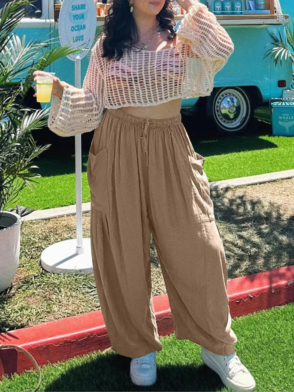 Person wearing a beige mesh top and wide-leg pants, holding a drink outdoors.