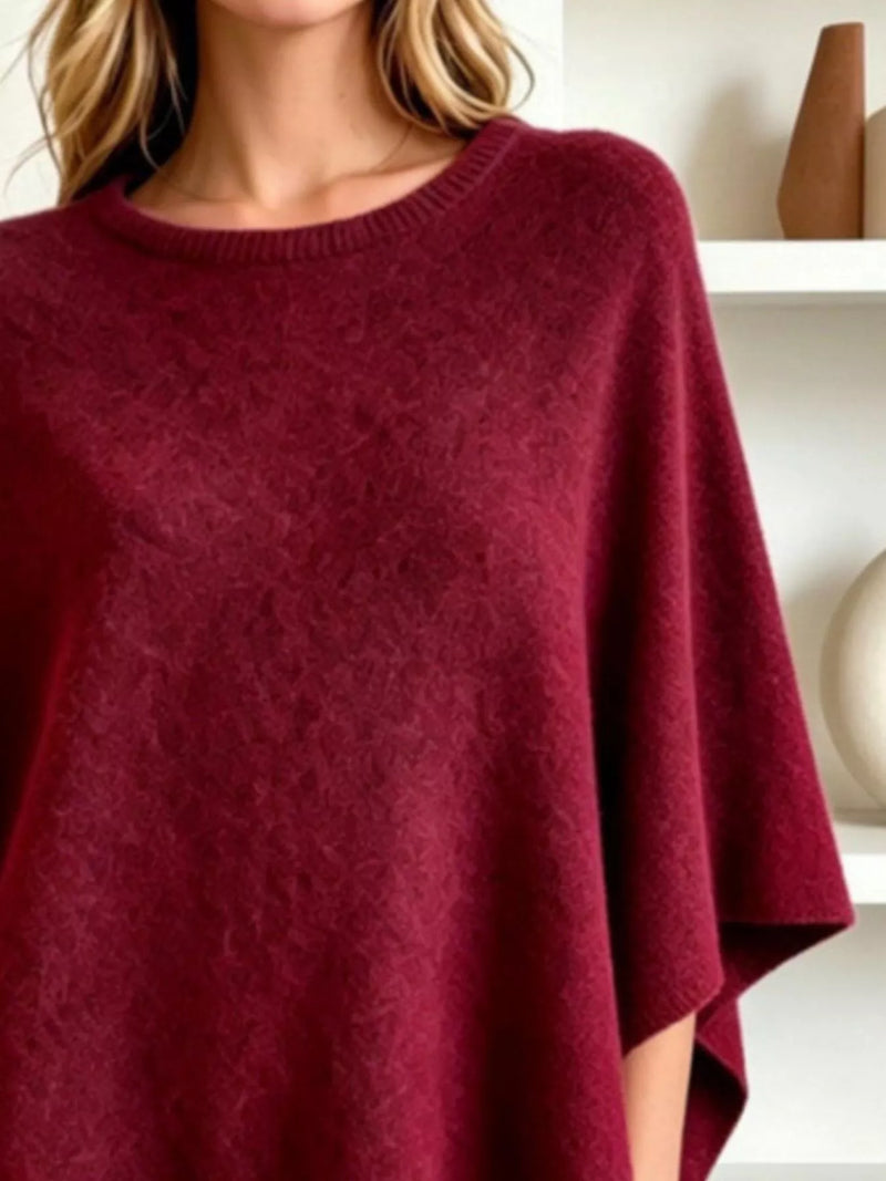 A close-up of a woman wearing a soft, textured knit poncho sweater in deep burgundy. The shot focuses on the rich fabric texture and the relaxed fit of the garment.