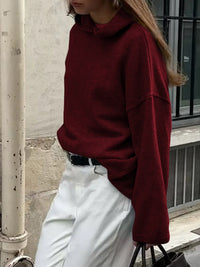 Person wearing a red sweater and white pants standing against a neutral wall.