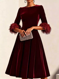 Burgundy velvet dress with feathered cuffs held by a hand with a clutch against a neutral background