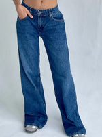 A person wearing loose, wide-leg blue denim jeans with a faded wash, shown from the waist down against a plain light gray studio background.