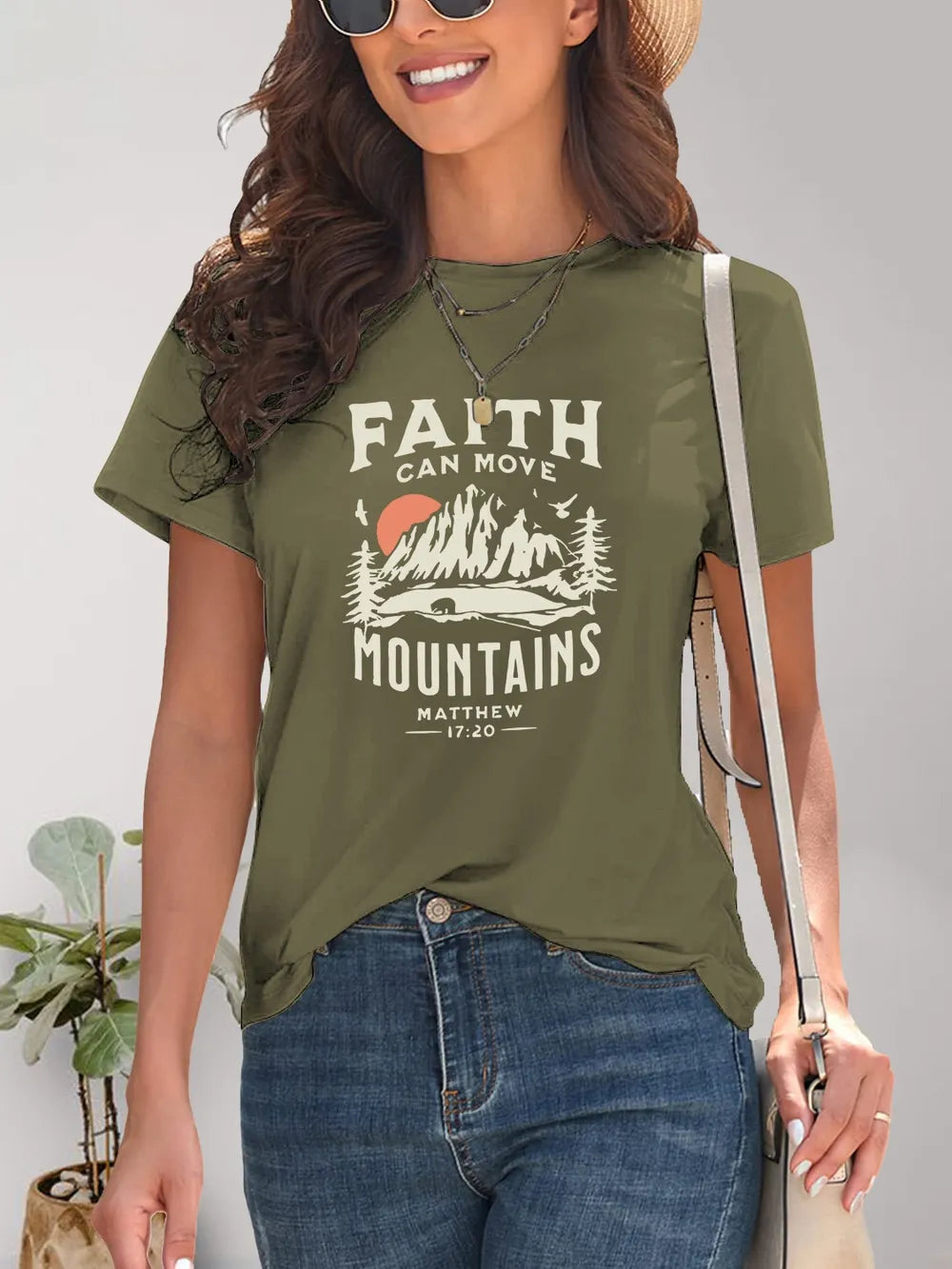 Woman wearing an olive green t-shirt with a mountain graphic and text, standing against a plain background.
