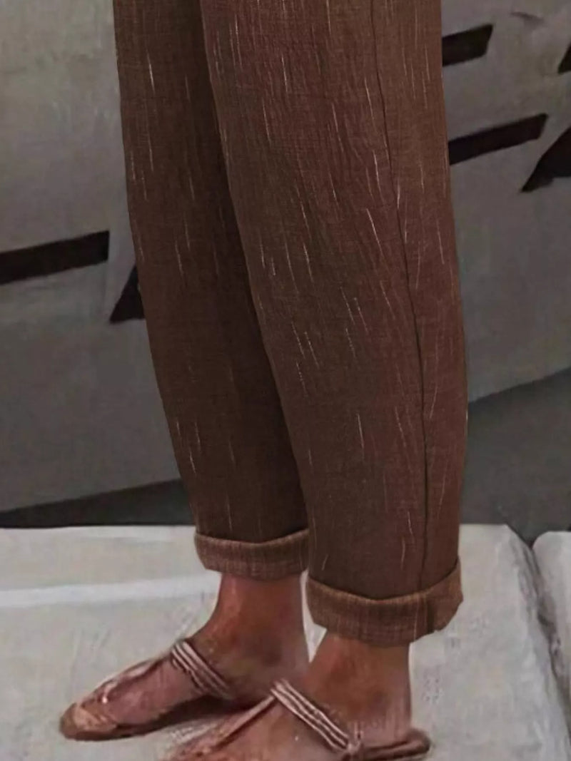 Close-up of a person's legs wearing brown textured cuffed pants and matching brown flat sandals with woven straps, standing on a light-colored surface.