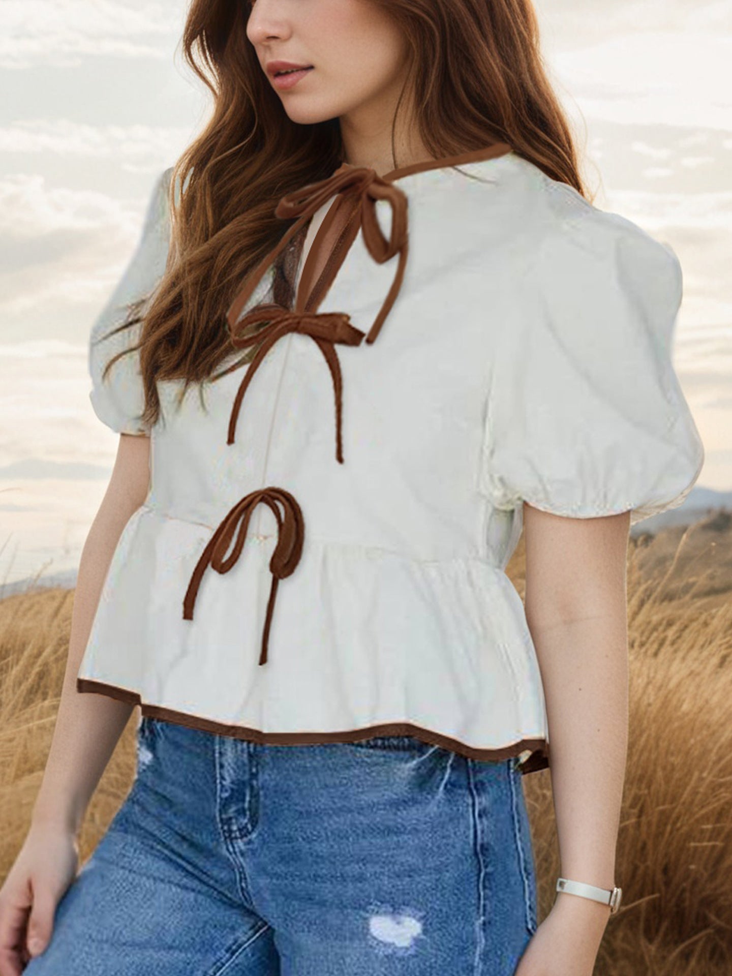 Woman wearing a white blouse with brown details and blue jeans in a natural setting.