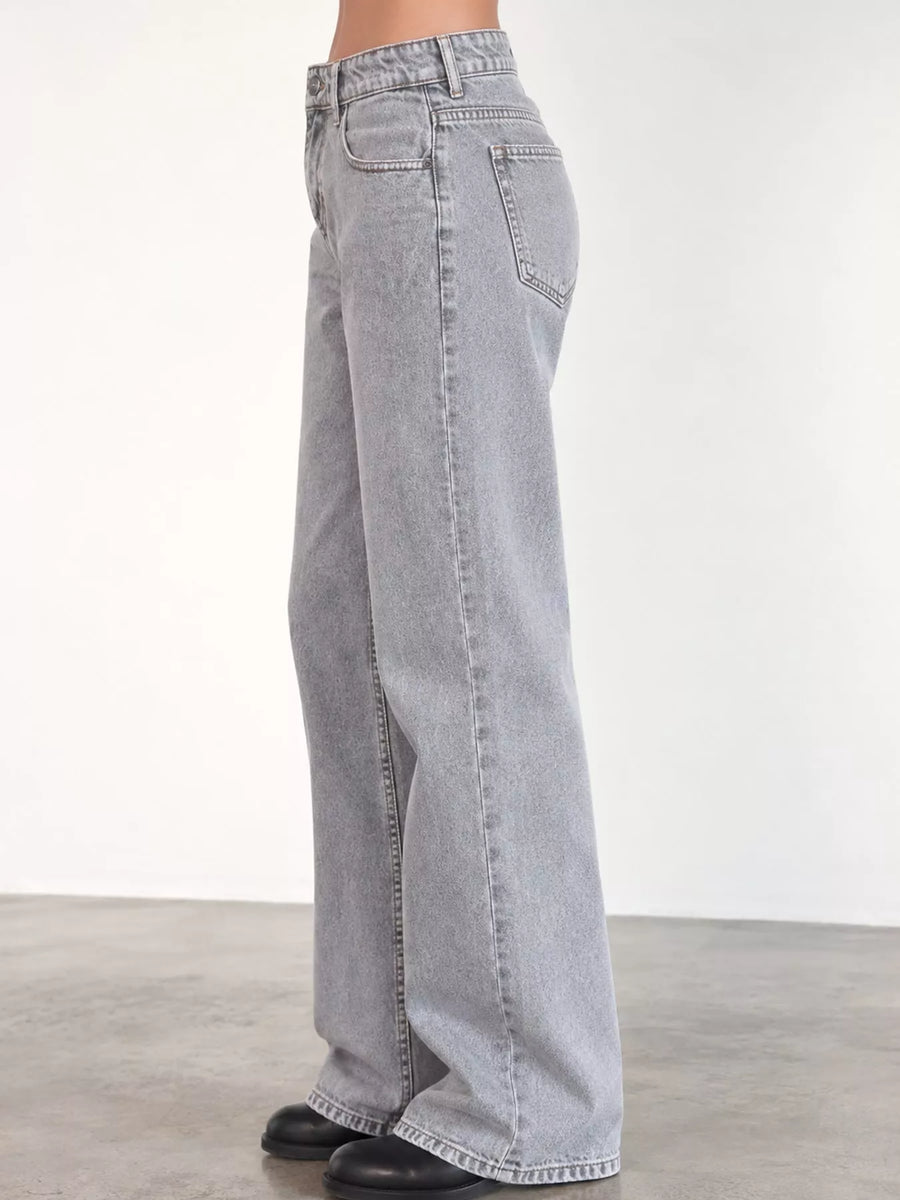 Light gray wide-leg denim jeans shown on a model from the side, paired with black shoes against a plain studio background.