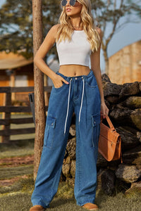 Woman wearing a white crop top and blue jeans, standing outdoors near a wooden post and stone wall.