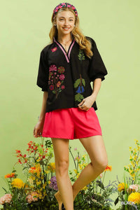 Woman wearing a black embroidered blouse and red shorts with flowers and greenery in the background