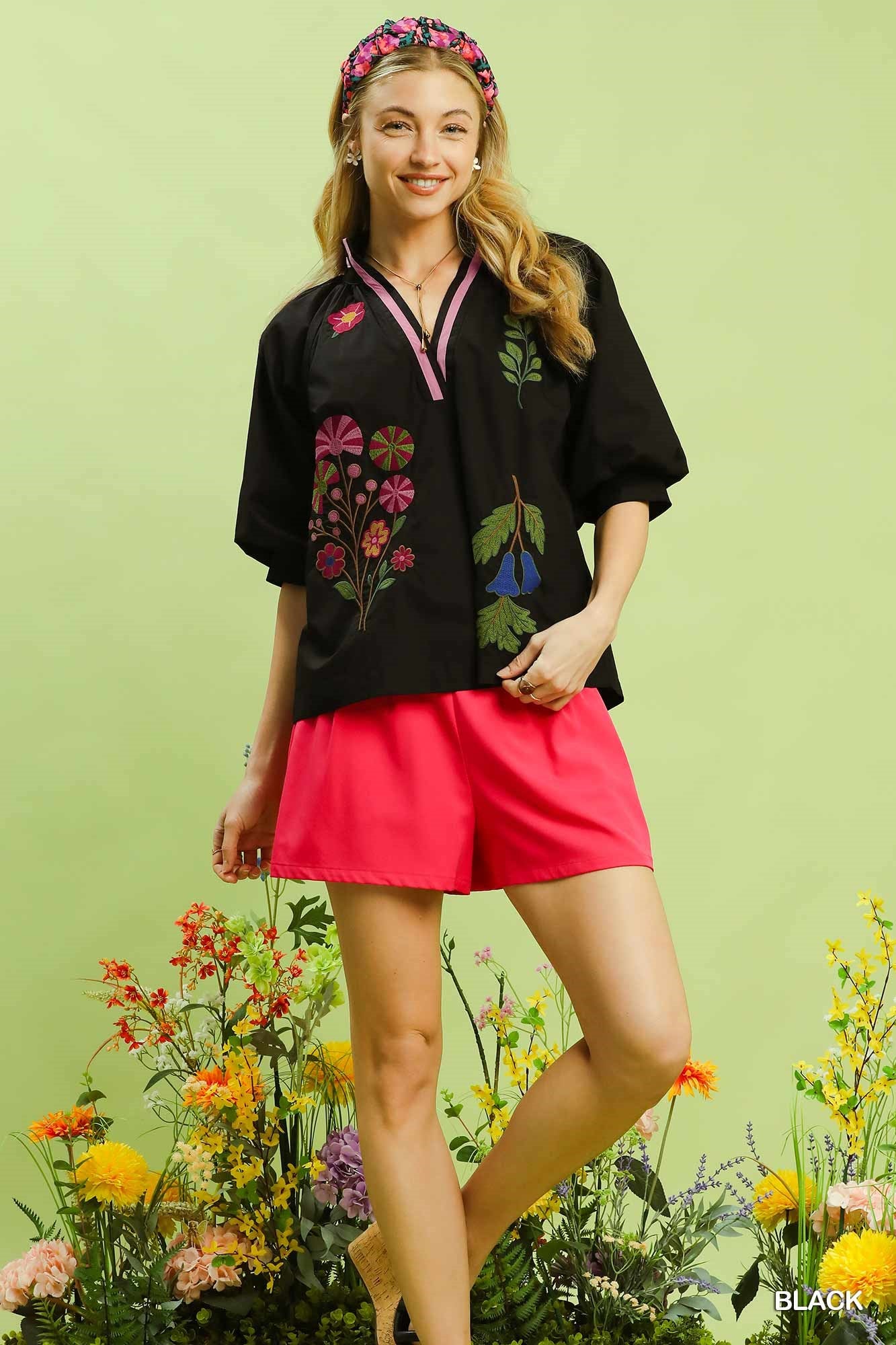 Woman wearing a black embroidered blouse and red shorts with flowers and greenery in the background