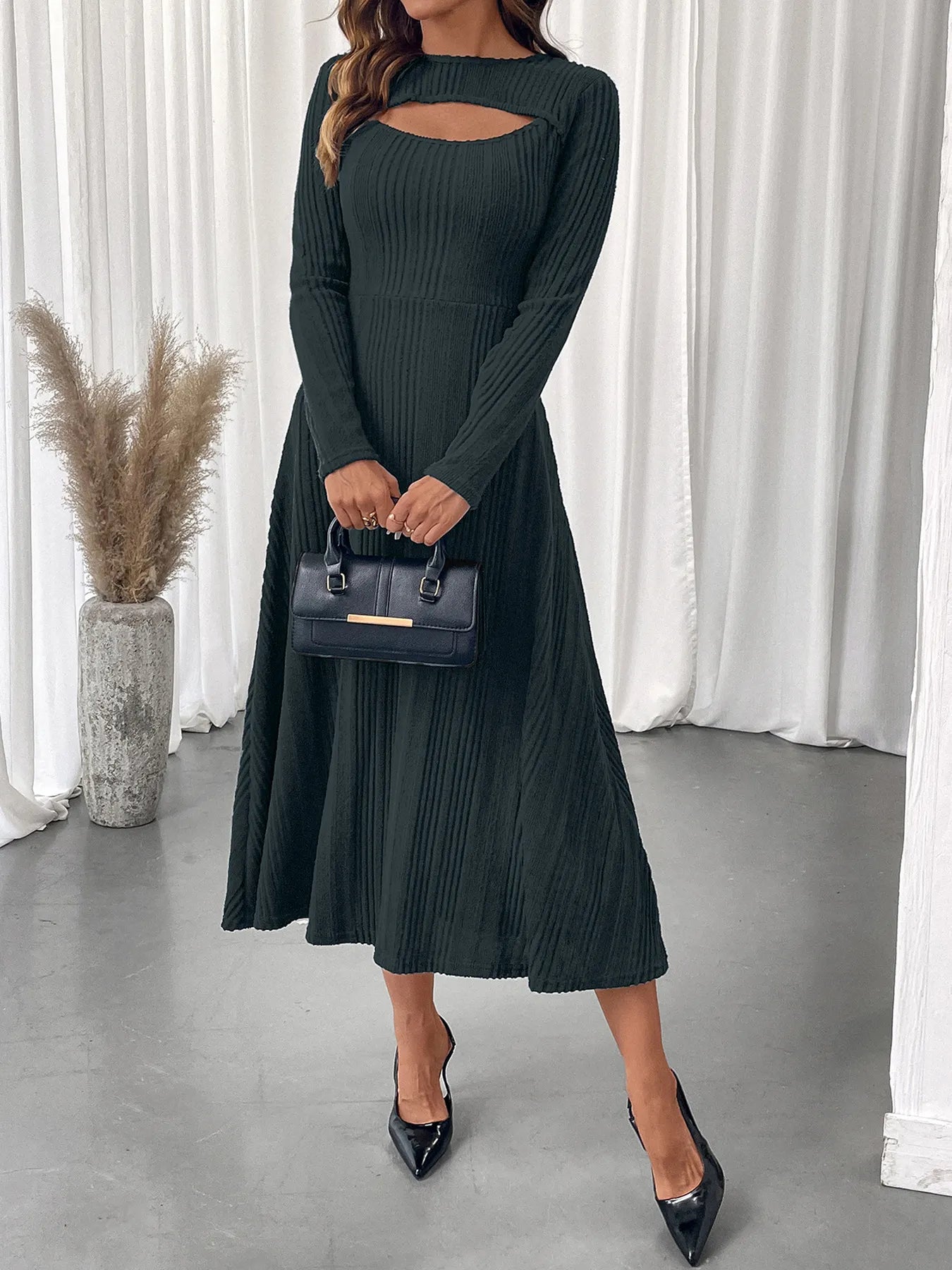 Woman wearing a dark green ribbed dress with cut-out design, holding a black handbag indoors.