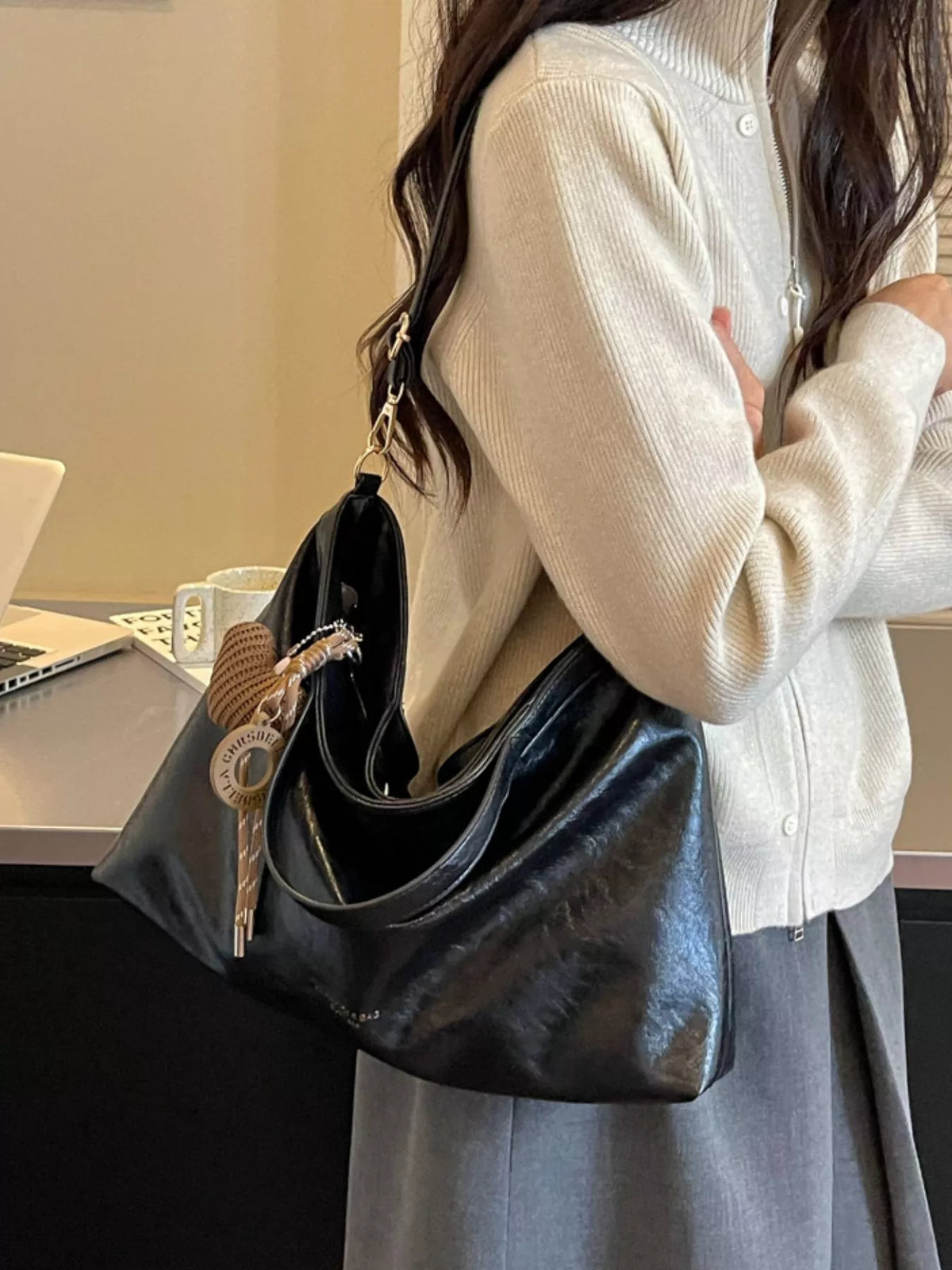 A person wearing a cream cardigan and gray skirt carries a large black slouchy shoulder bag with a decorative key charm in an indoor setting.