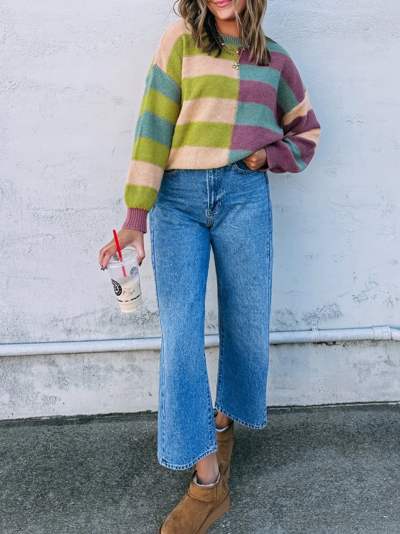 Person wearing a colorful striped sweater and blue jeans, holding a drink against a white wall.
