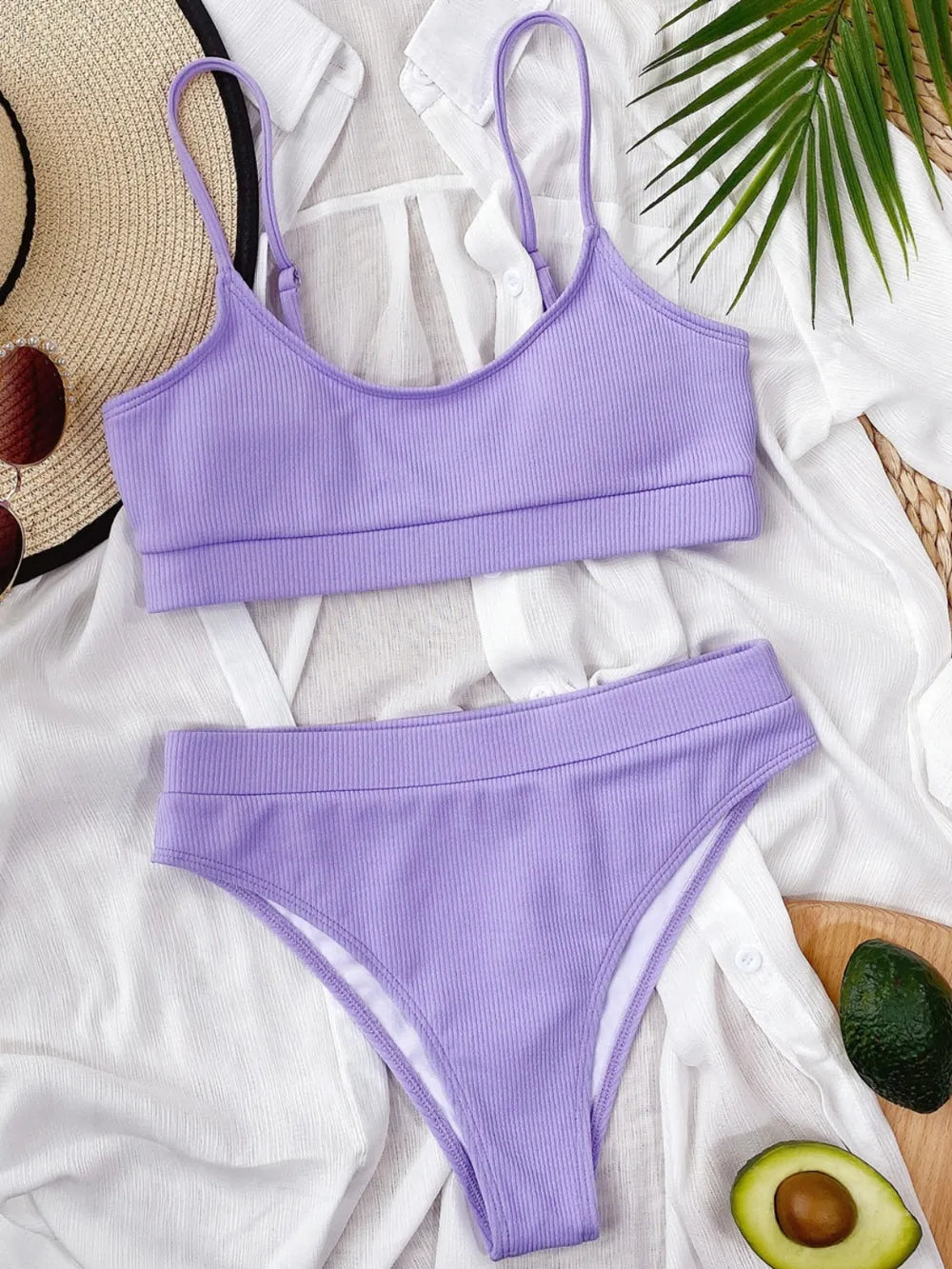 Purple bikini set on a white surface with a hat and avocado in the background