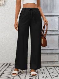 Person wearing black wide-leg pants with a white top and sandals, holding a brown handbag against a light gray wall.