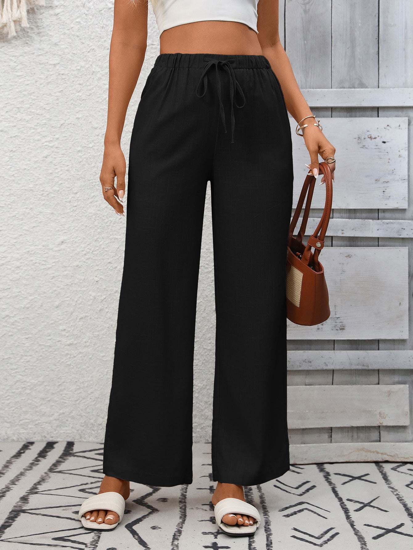 Person wearing black wide-leg pants with a white top and sandals, holding a brown handbag against a light gray wall.