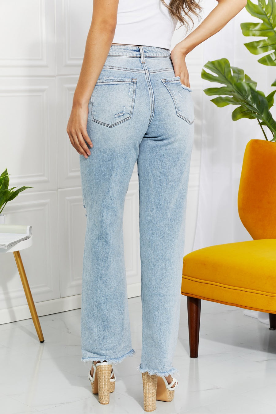 Person wearing light blue jeans standing in a room with a white wall, yellow chair, and green plant.