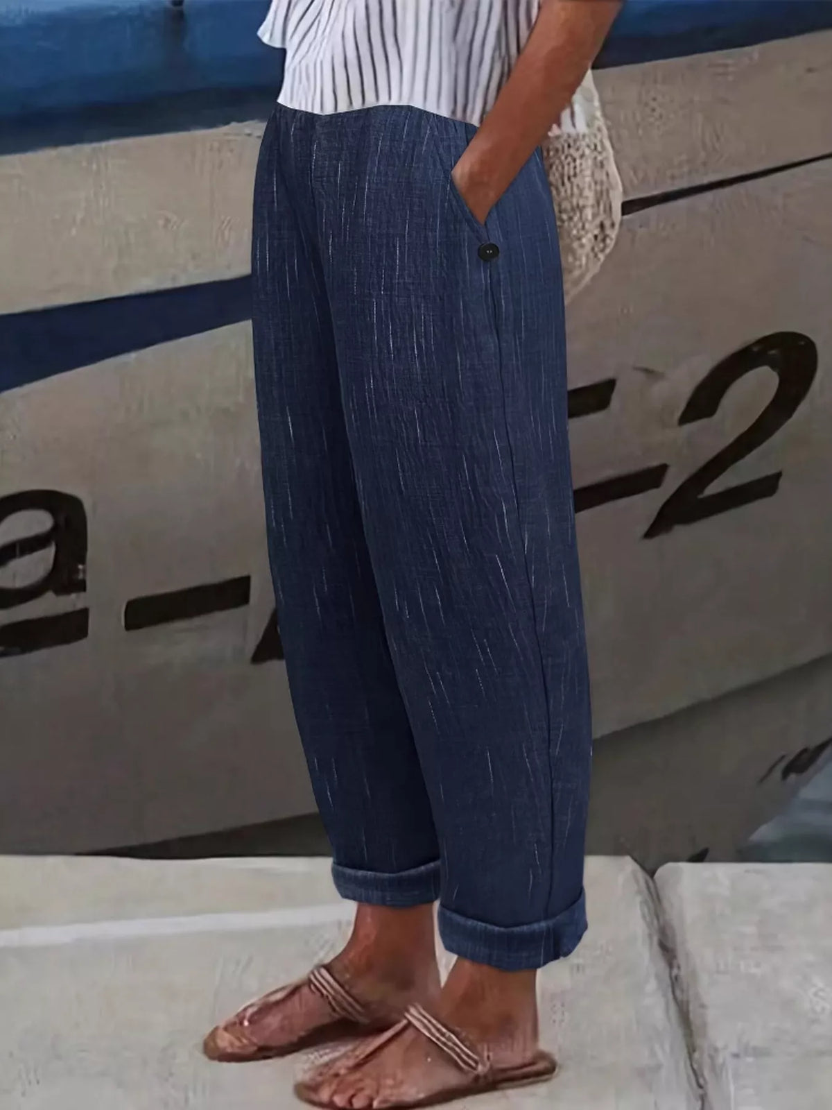 A person wearing loose, dark blue textured pants with rolled cuffs, paired with a white striped top and brown sandals, standing outdoors against a boat hull.