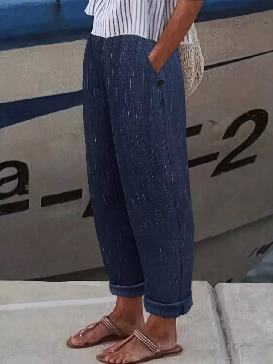 A person wearing loose, dark blue textured pants with rolled cuffs, paired with a white striped top and brown sandals, standing outdoors against a boat hull.