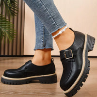 Black leather shoes with buckles worn with blue jeans on a wooden floor.