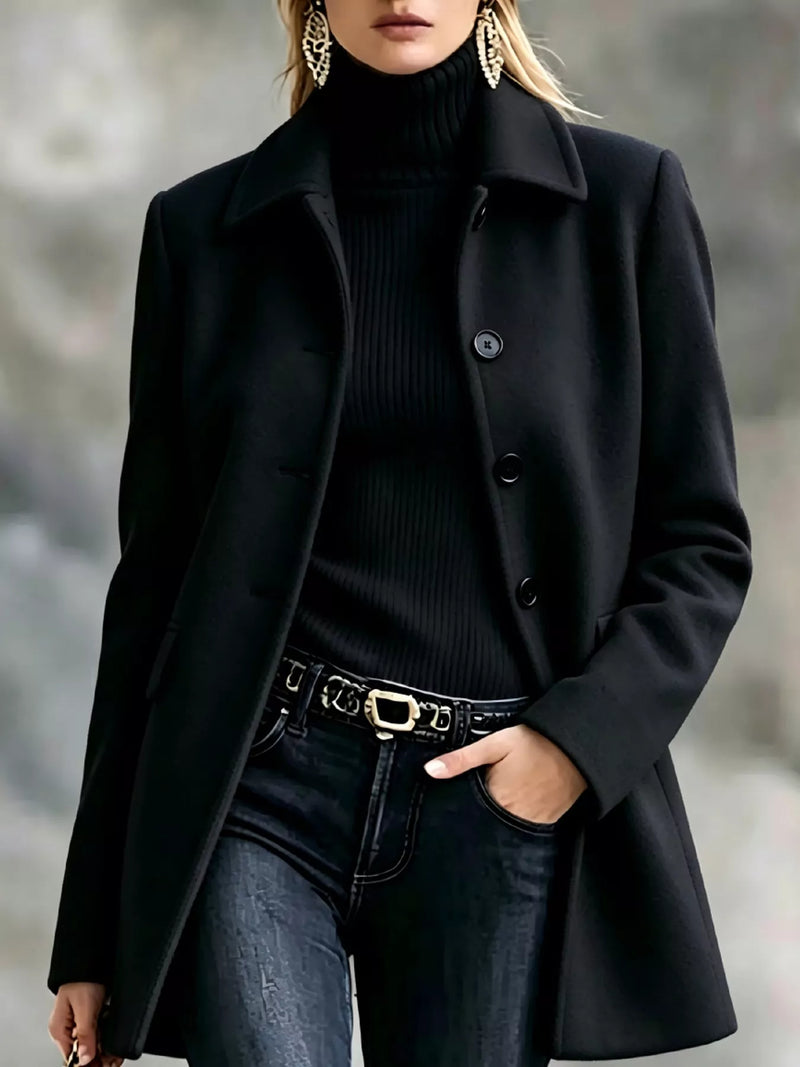 A woman wearing a black mid-length coat over a black ribbed turtleneck sweater, paired with dark jeans and a black belt with a gold buckle, photographed outdoors against a blurred gray background.