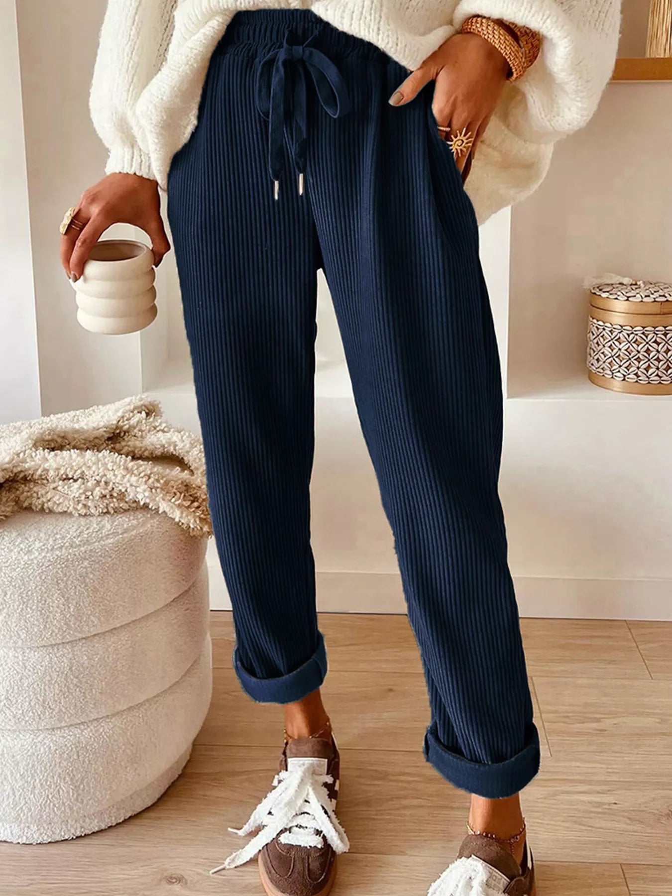 Person wearing navy ribbed pants with a white sweater and brown shoes, holding a small white container.