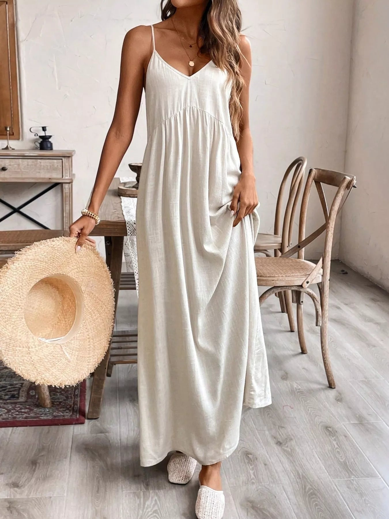 Woman in a long white dress holding a straw hat in a room with wooden furniture.