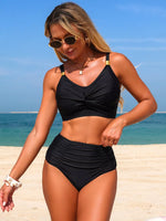 Woman in a black bikini standing on a beach with clear blue sky and water.