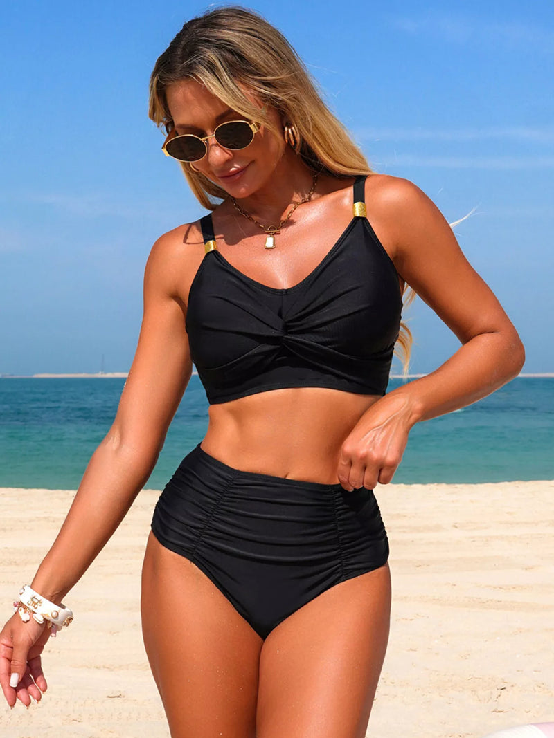 Woman in a black bikini standing on a beach with clear blue sky and water.