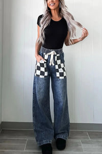 Woman wearing wide-leg jeans with a checkered pocket and black top against a white wall.