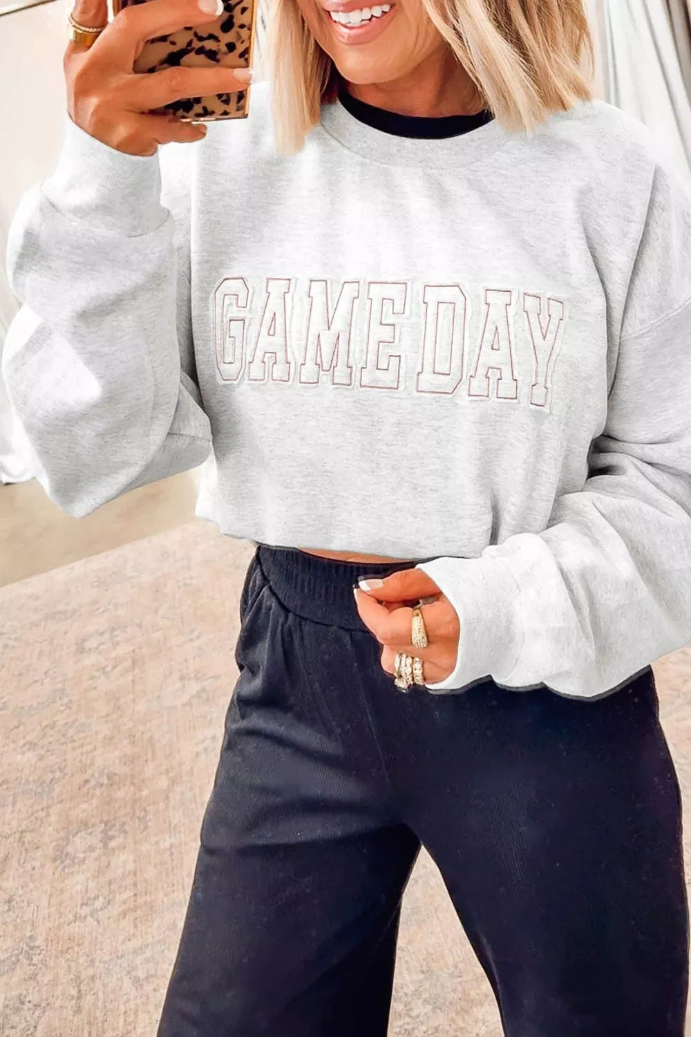 A woman takes a mirror selfie wearing a light gray cropped sweatshirt with 'GAME DAY' printed across the chest in a double-outline font, paired with dark pants.