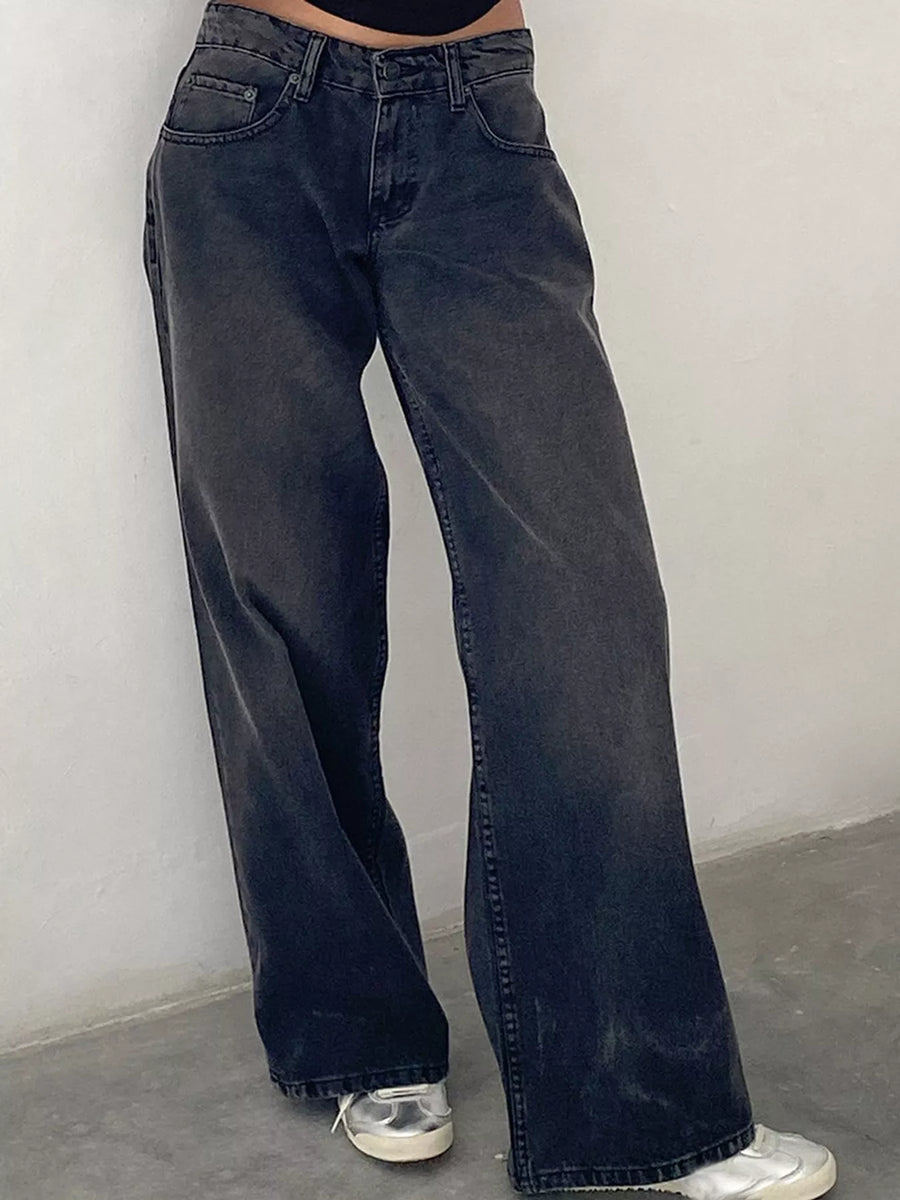 A person wearing dark washed wide-leg denim jeans with silver sneakers, standing against a plain light gray wall.