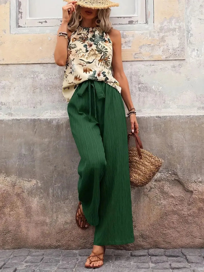 Woman wearing a floral top and green pants against a textured wall.