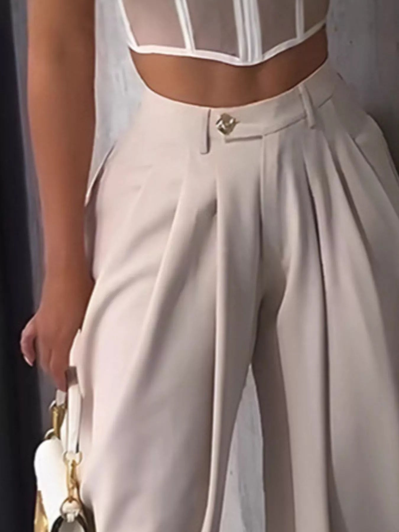 Person wearing a white blouse with sheer panels and beige high-waisted pants, holding a white handbag.
